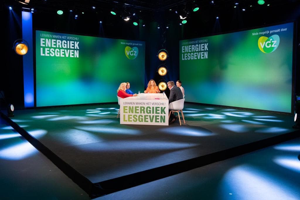 Studio met groene lichten en de tekst 'enegeriek lesgeven' met mensen zittend aan tafel