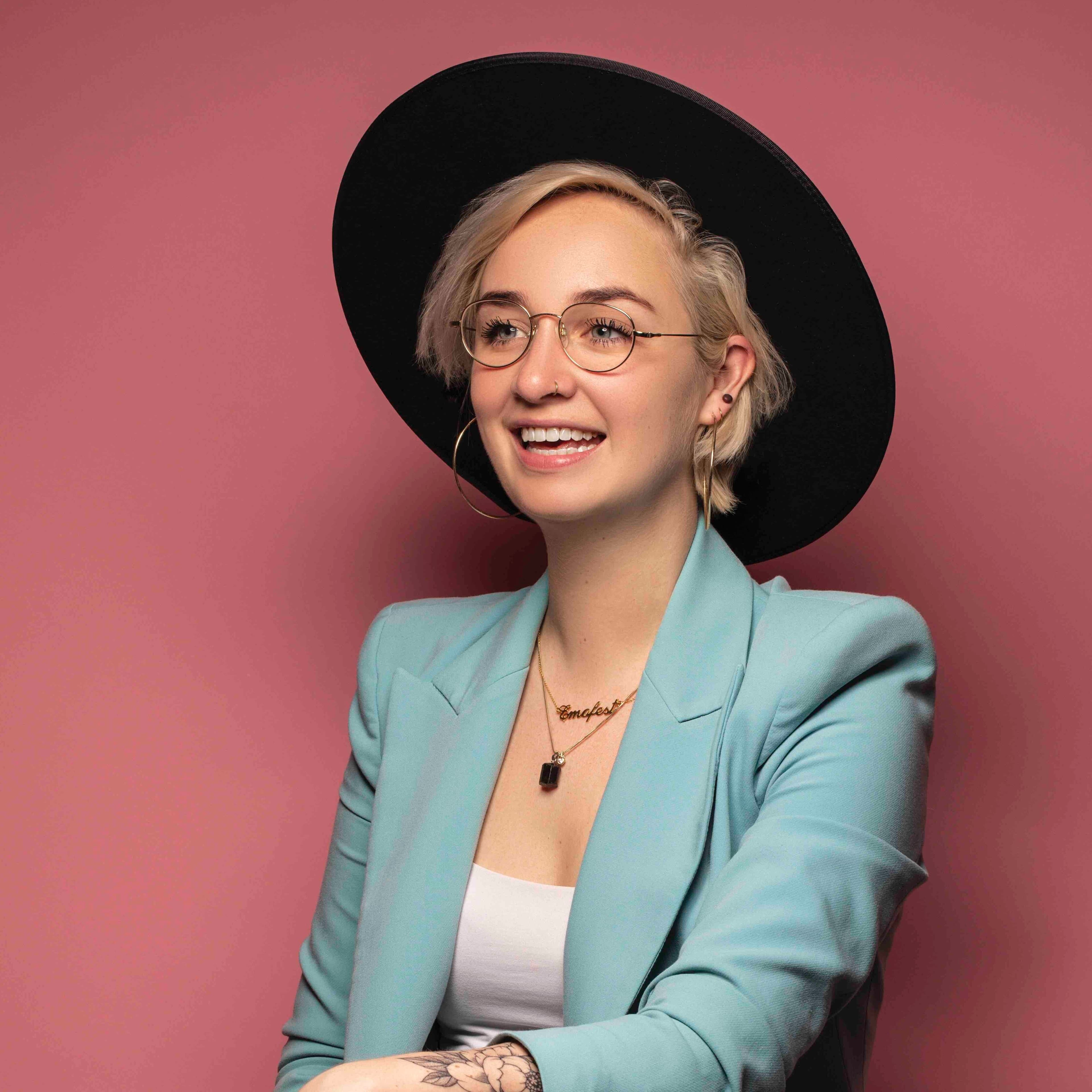 Danielle Jiskoot lachend met zwarte hoed en blauwe blazer bij roze achtergrond 