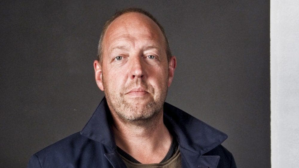 Harry Piekema poserend in blauwe jas bij grijze achtergrond