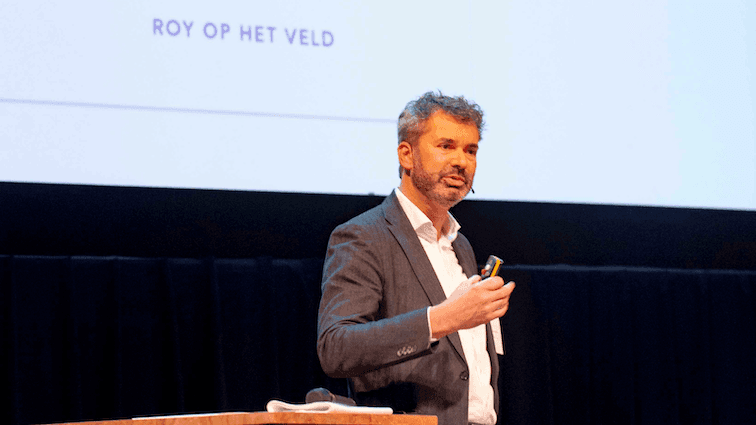Roy op het Veld aan het presenteren in grijs pak en beweegt met hand