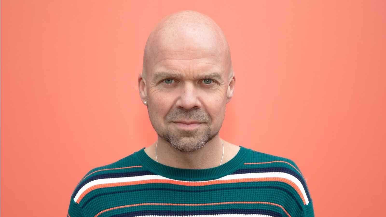 Andre Heuvelman poserend in groene sweater met strepen bij oranje achtergrond