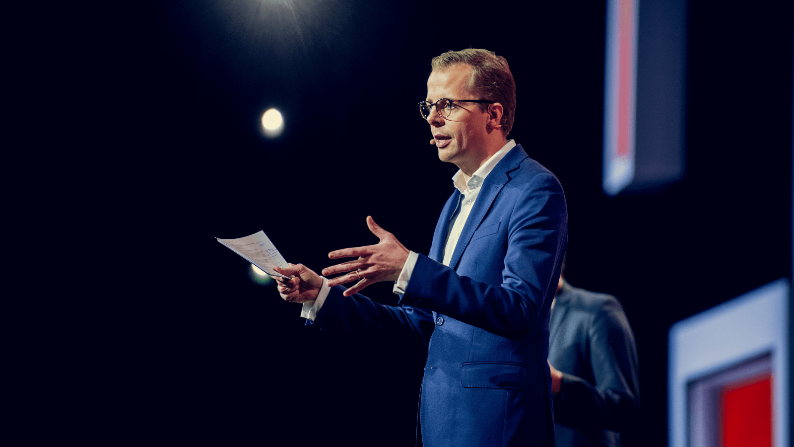 Jeroen Baardemans aan het presenteren bewegend met handen in blauw pak op podium