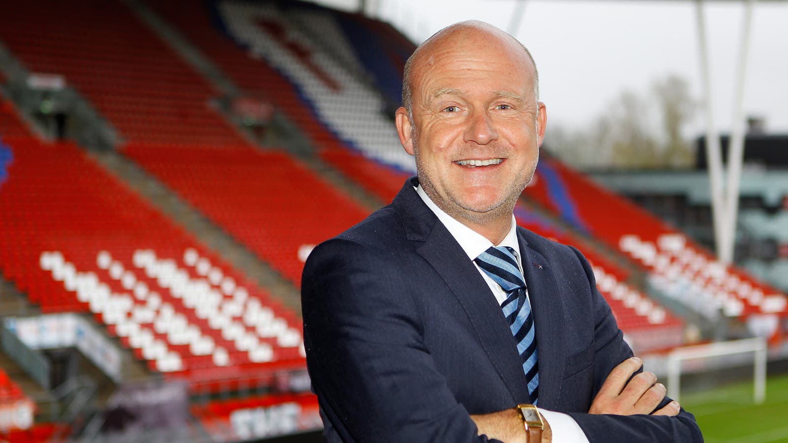 Wilco van Schaik met armen over elkaar glimlachend in blauw pak in een voetbalstadion