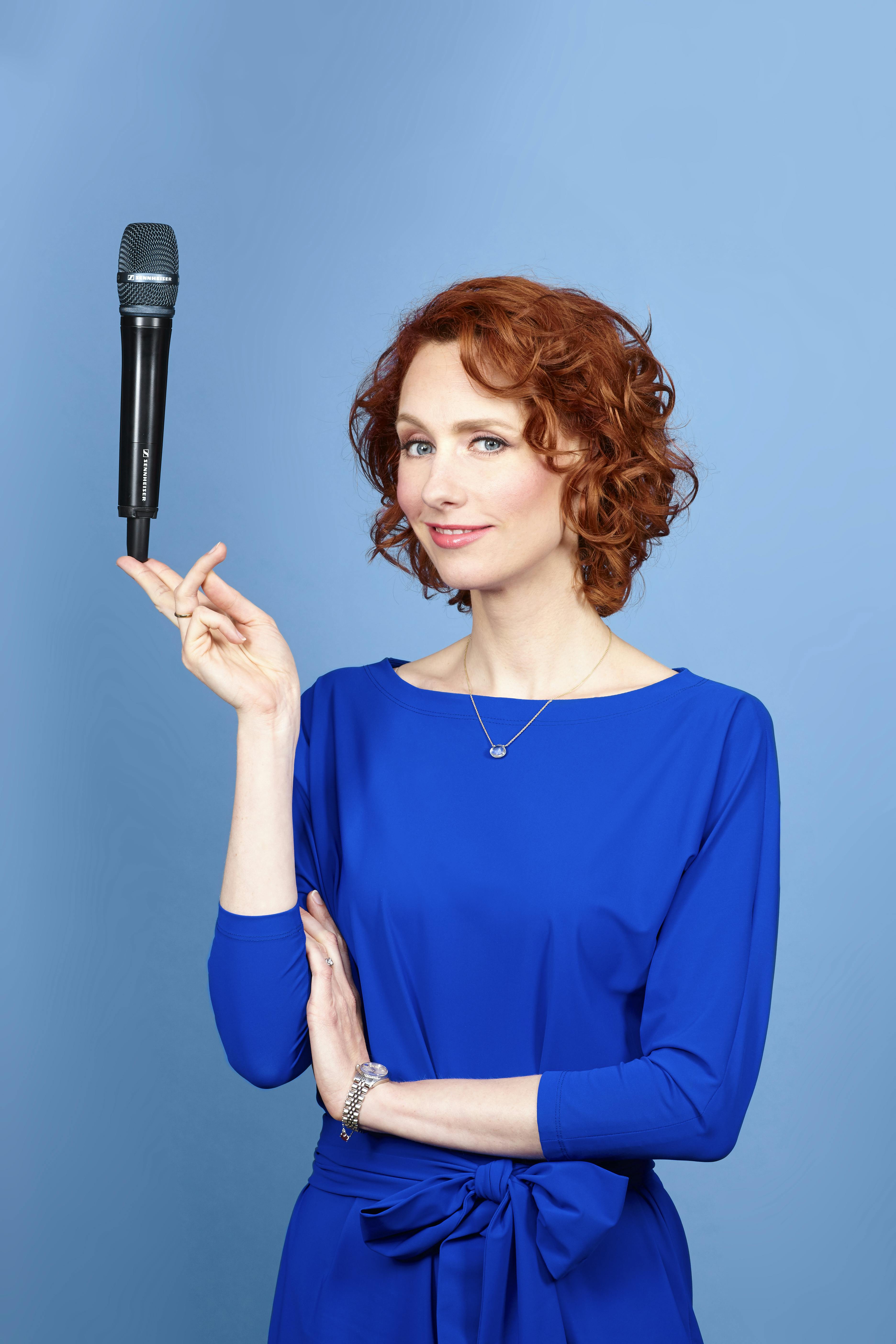 Eva Brouwer glimlachend in blauwe jurk met microfoon poserend op twee vingers bij blauwe achtergrond