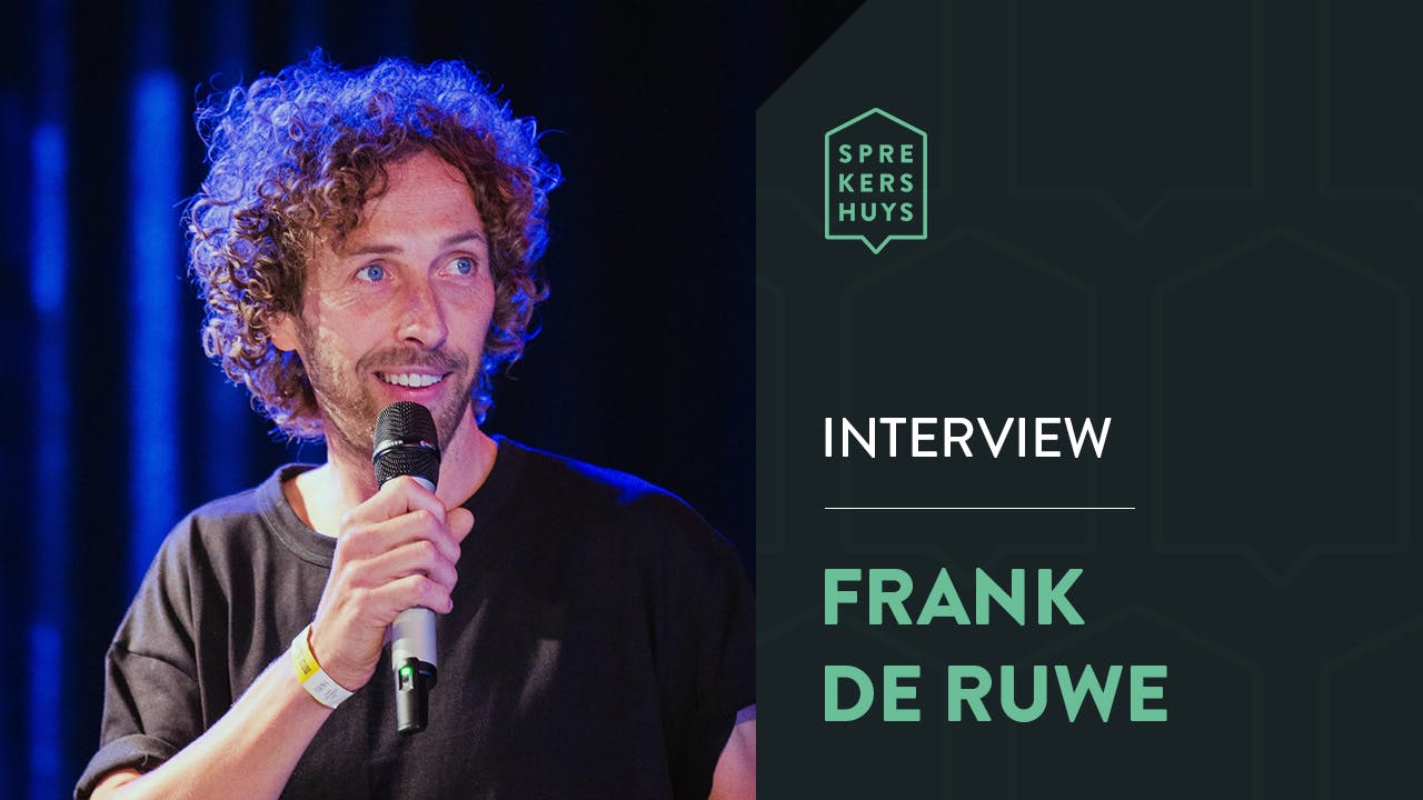 Frank de Ruwe met microfoon bij de mond en blauwe gloed achter zich met de tekst 'Interview Frank de Ruwe'