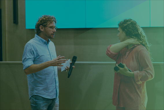 Ewout Genemans aan het praten met vrouw en beweegt handen met microfoon in blauwe pak met groene gloed over foto heen
