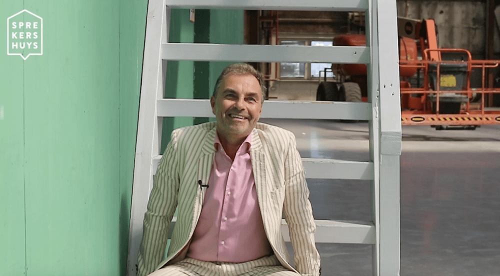 Stephan Fellinger zittend op witte trap aan het glimlachen in beige pak en roze blouse