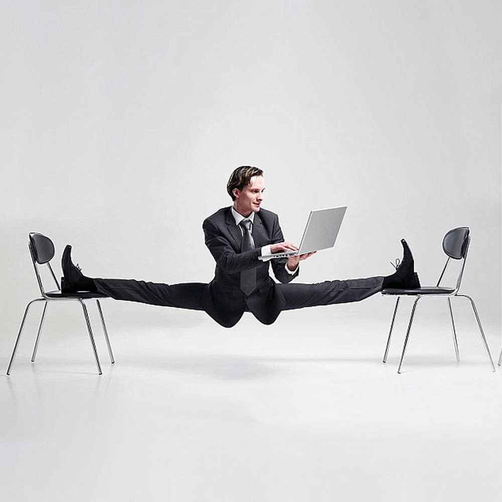 Man in zwart pak in de split met zijn benen op twee stoelen kijkend naar laptop in zijn handen