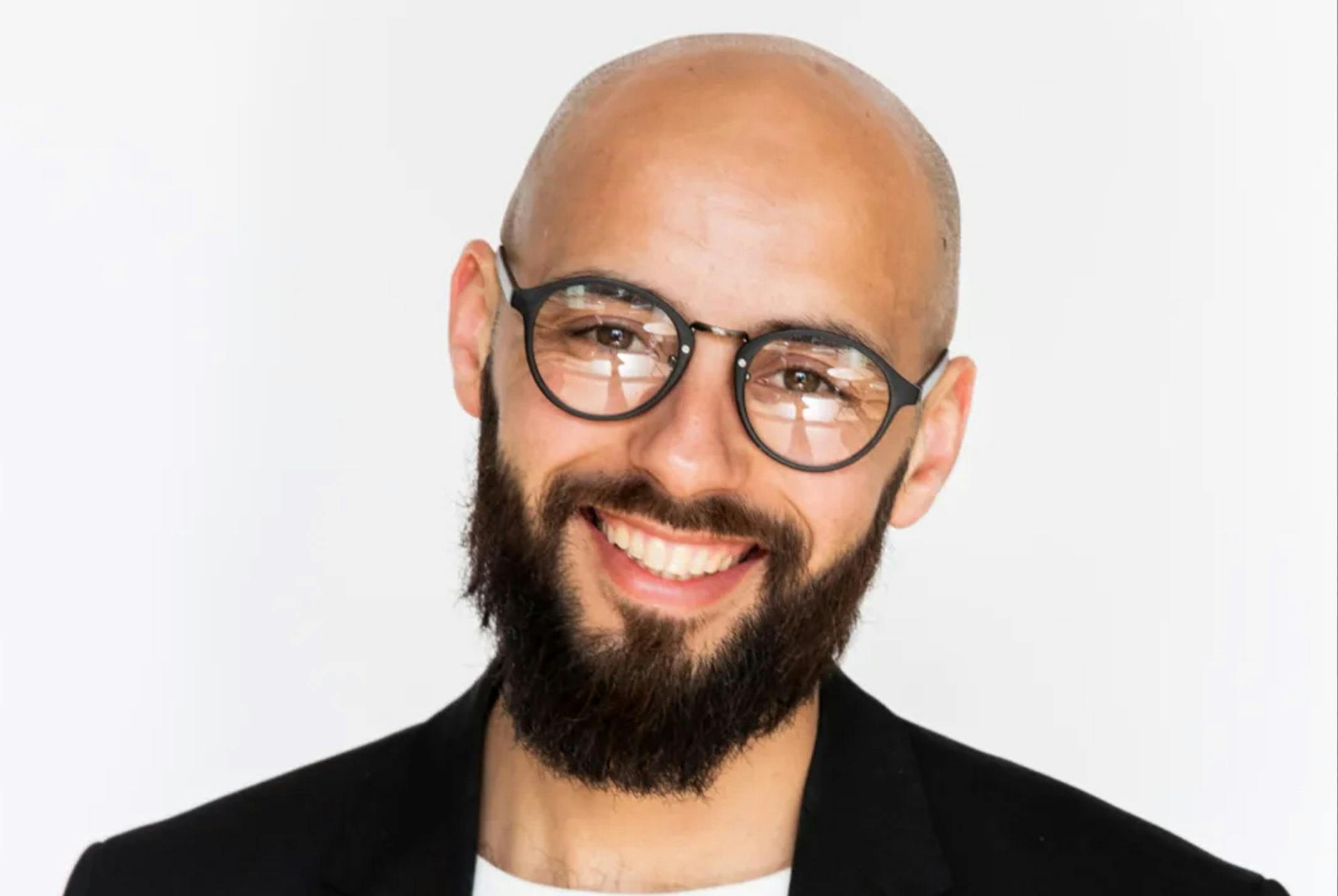 Karim Amghar glimlachend in zwart pak bij witte achtergrond