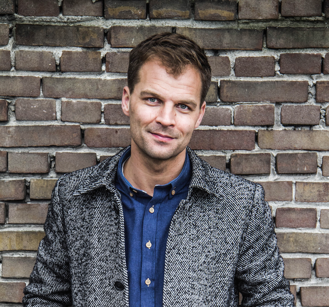 Daan Nieber poserend buiten bij stenen muur met grijze jas en blauwe blouse