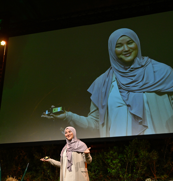 Houda Loukili voor groot scherm in zaal met handen omhoog