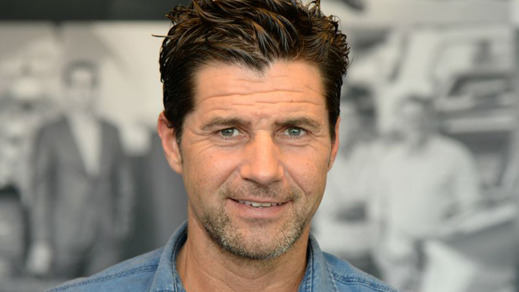 Man met een kleine baard en in het blauw gekleed achter een zwart witte achtergrond