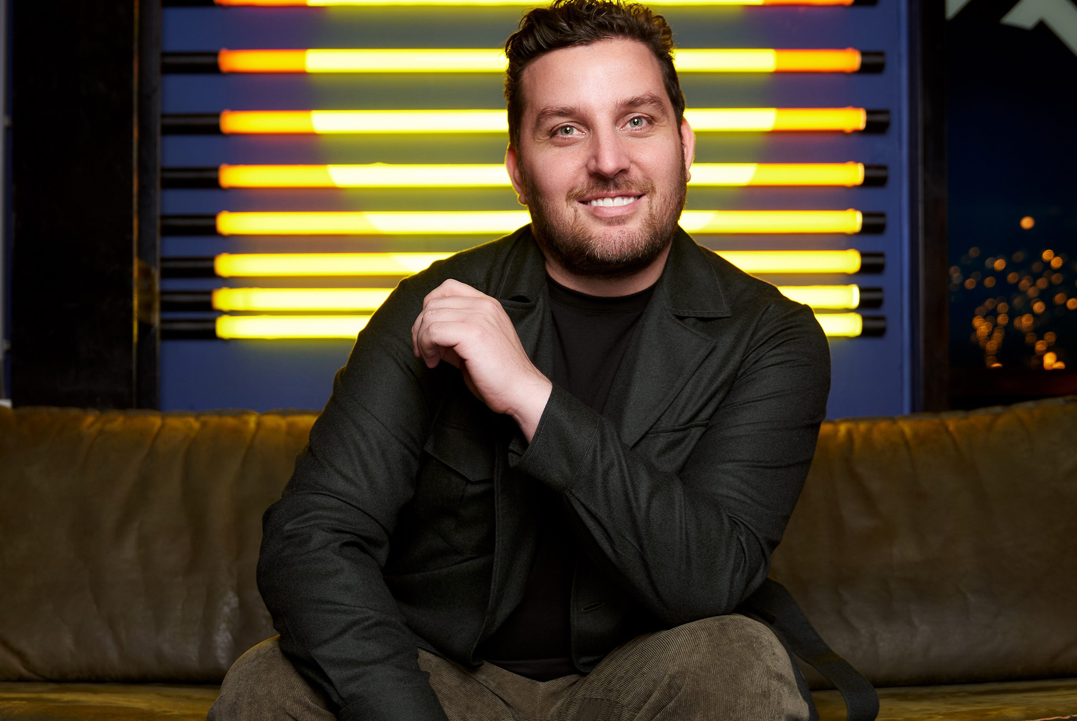 Ron Simpson poserend in zwarte jasje zittend op een bank met gele lampen achter zich