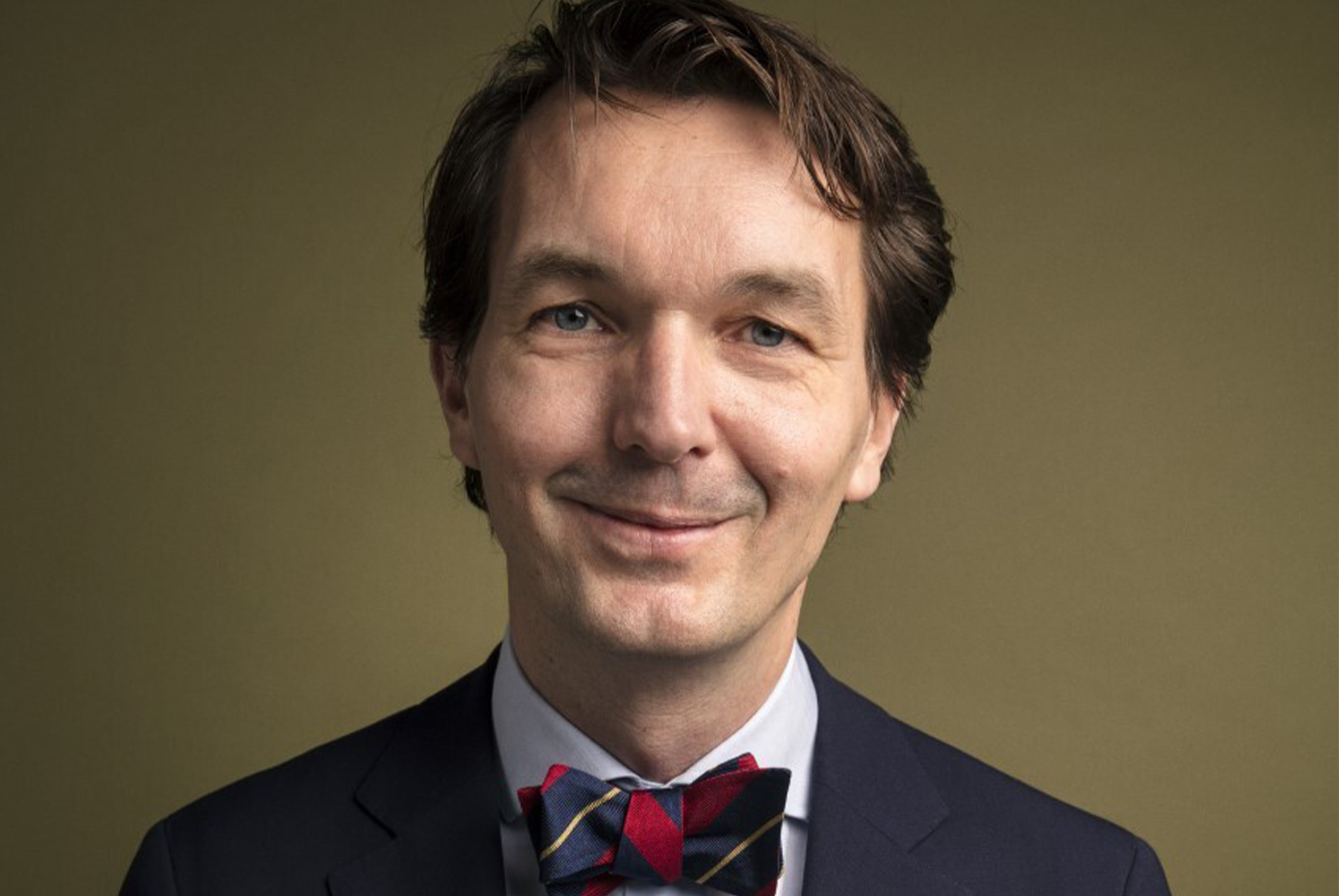 Paul van Riessen glimlachend in zwart pak bij bruine achtergrond