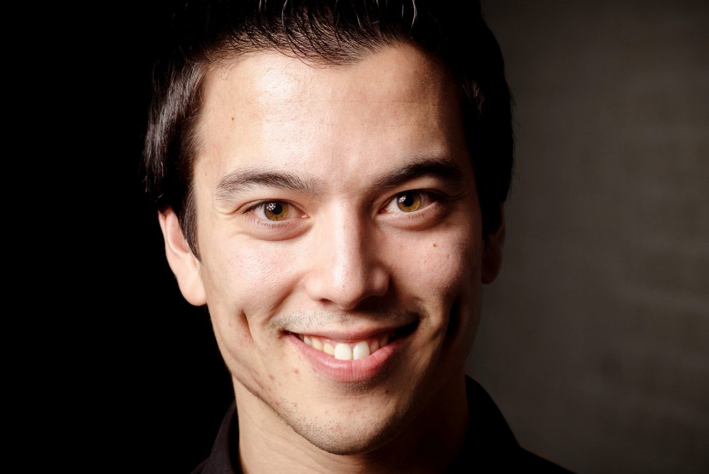 Andries Tunru glimlachend in zwarte blazer bij grijze achtergrond