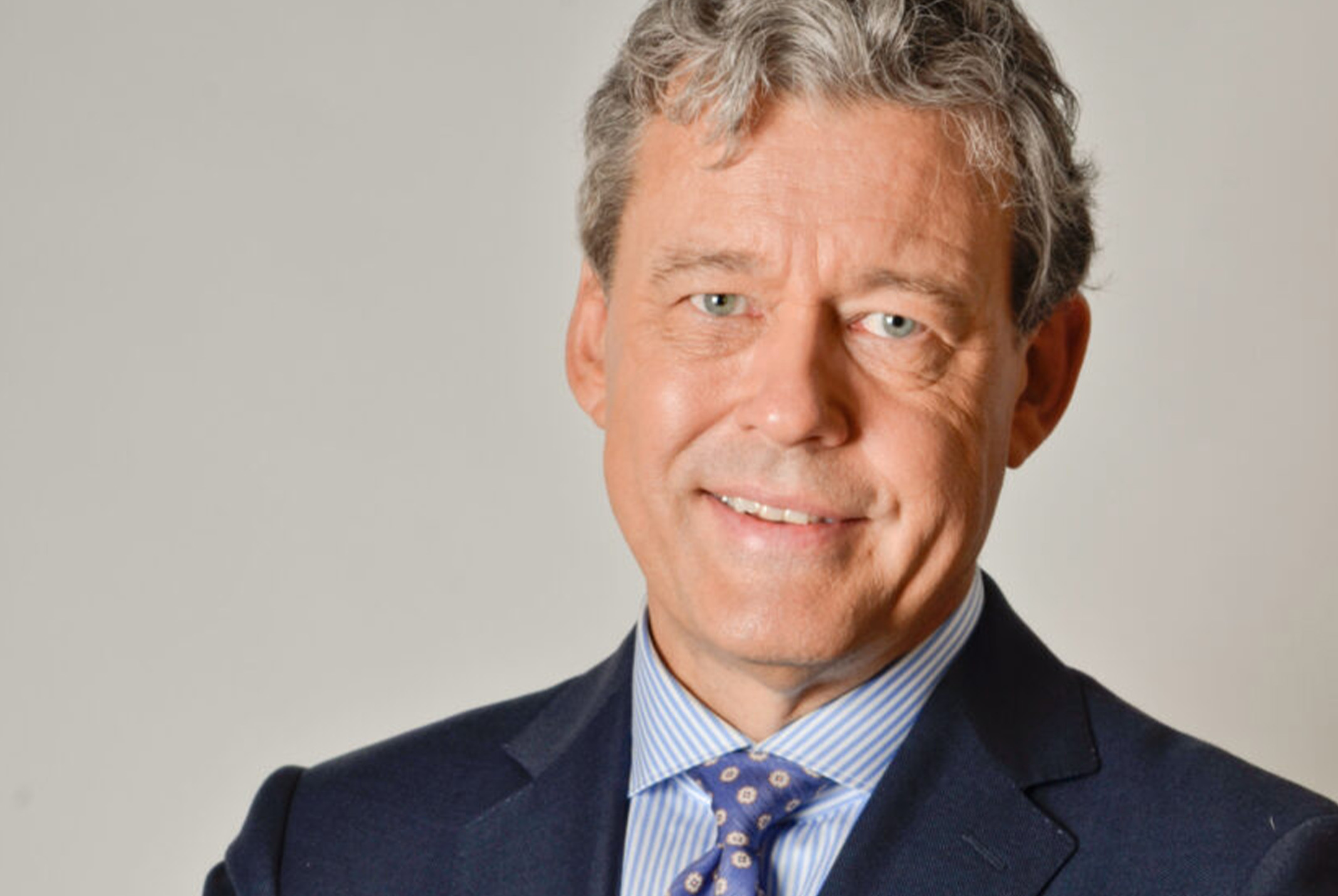 Charles Groenhuijsen glimlachend in pak bij beige achtergrond