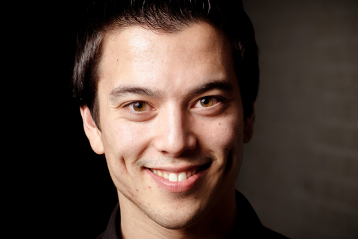 Andries Tunru glimlachend in zwarte blazer bij grijze achtergrond