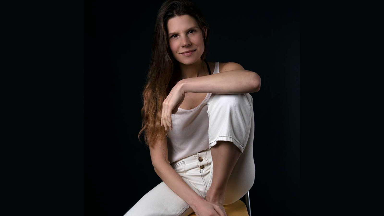 Roanne van Voost zittend op stoel met een been op de stoel en arm op been in witte kleding glimlachend bij zwarte achtergrond