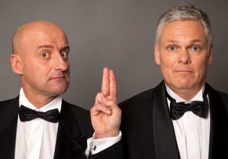 Joep van Deudekom en Rob Urgert in zwart pak die twee vingers opsteken bij grijze achtergrond