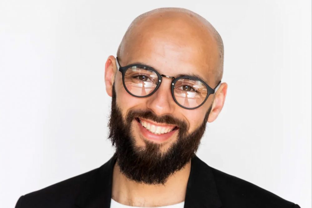 Karim Amghar glimlachend in zwart pak bij witte achtergrond