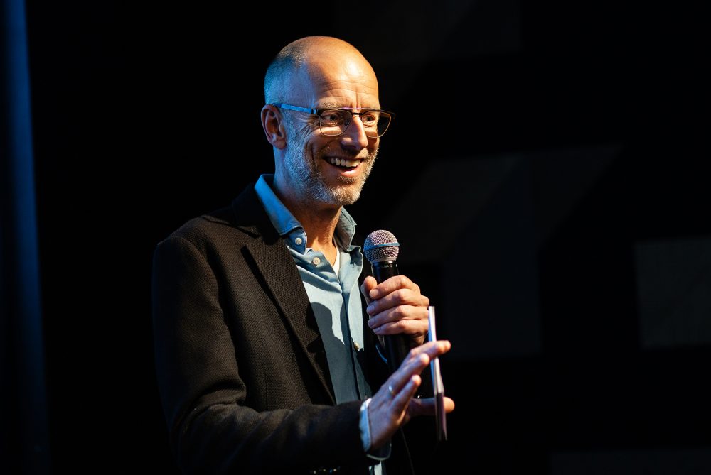 Sander de Heer lachend met microfoon in hand in zwarte blazer bij zwarte achtergrond