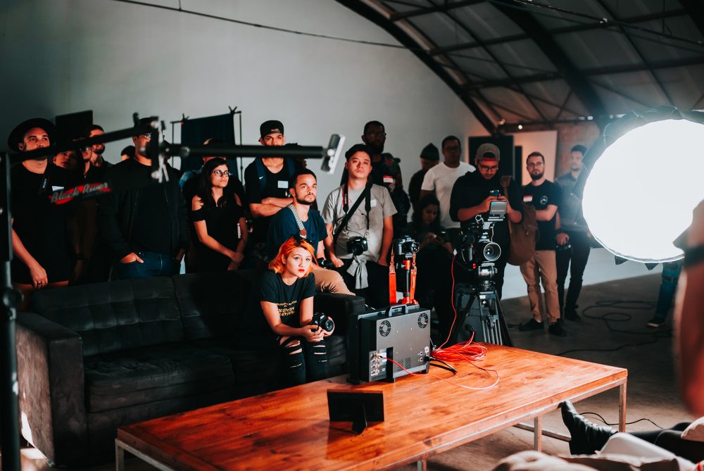Veel camera- en geluidmensen met apparatuur, een grote lamp en een houten tafel