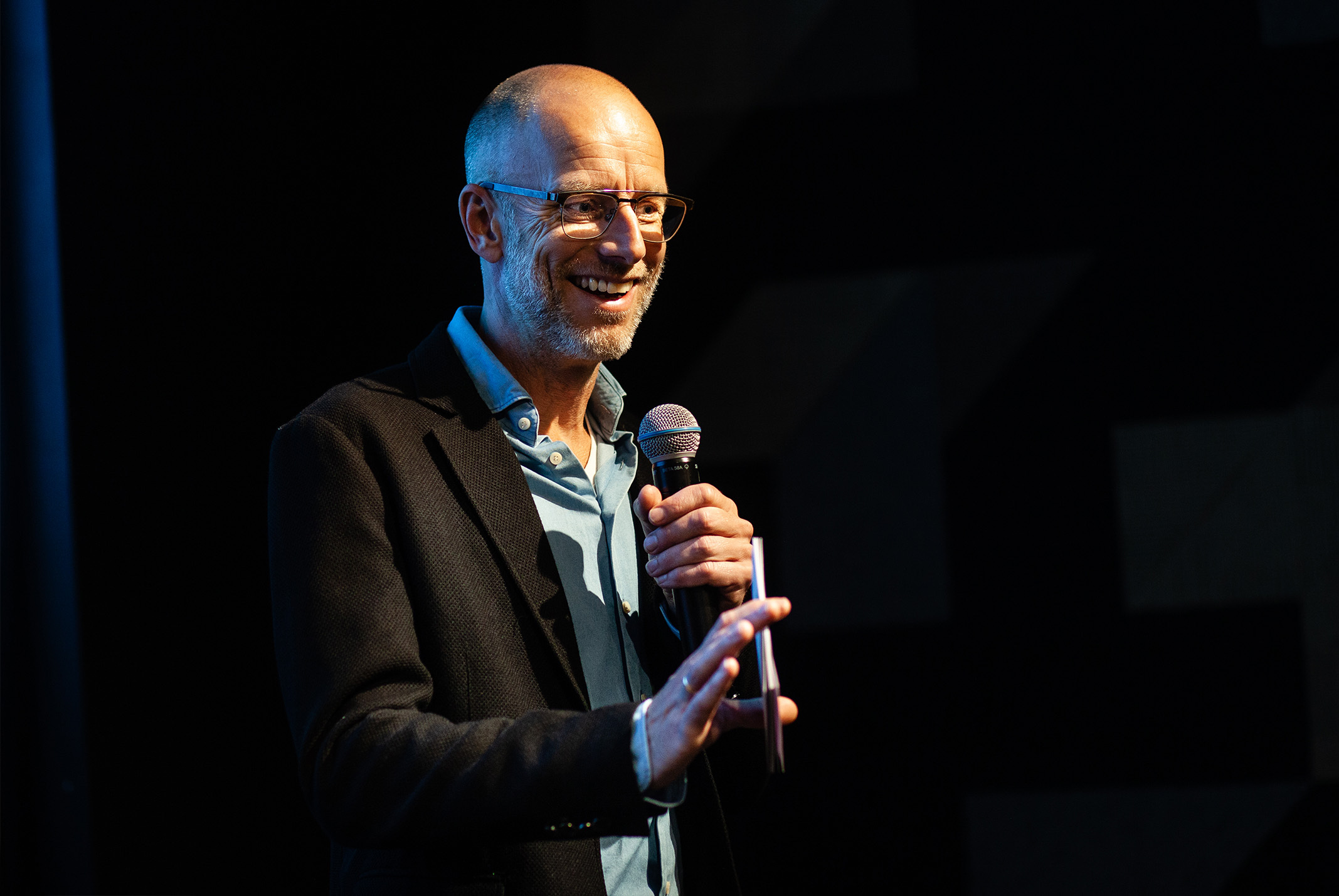 Sander de Heer lachend met microfoon in hand in zwarte blazer bij zwarte achtergrond