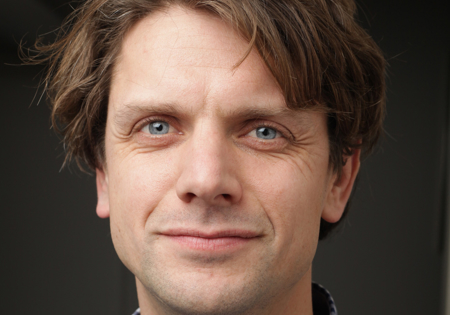 Jaap Vriend poserend in wit en blauwe geruiten blouse bij grijze achtergrond