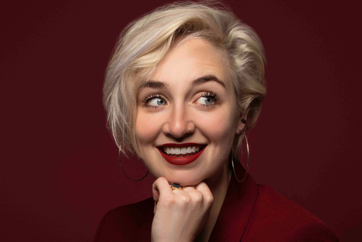 Danielle Jiskoot lachend kijkend naar zijkant met hand bij kin in rode blazer bij rode achtergrond