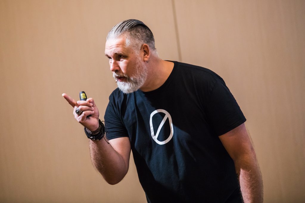 Edwin wijst met vingers ergens naartoe. Hij draagt een zwart shirt. Beige achtergrond