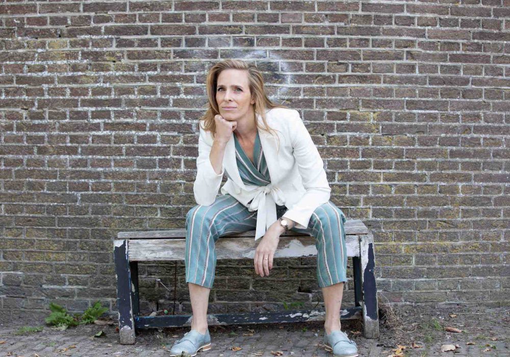 Kim Jansen poserend met hand onder haar kin zittend op een houten bankje buiten bij stenen muur