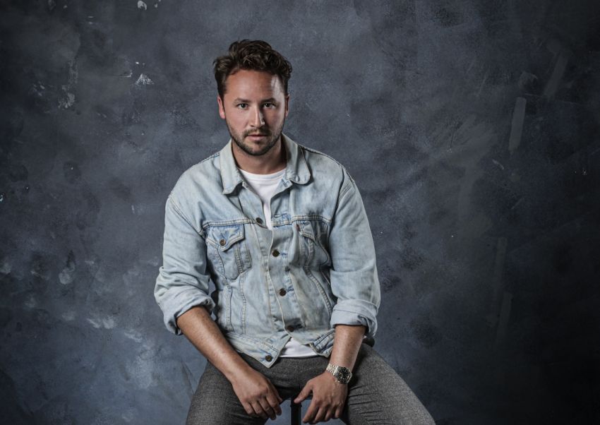 Daan Ubachs poserend zittend op stoel met spijkerjasje bij grijze achtergrond