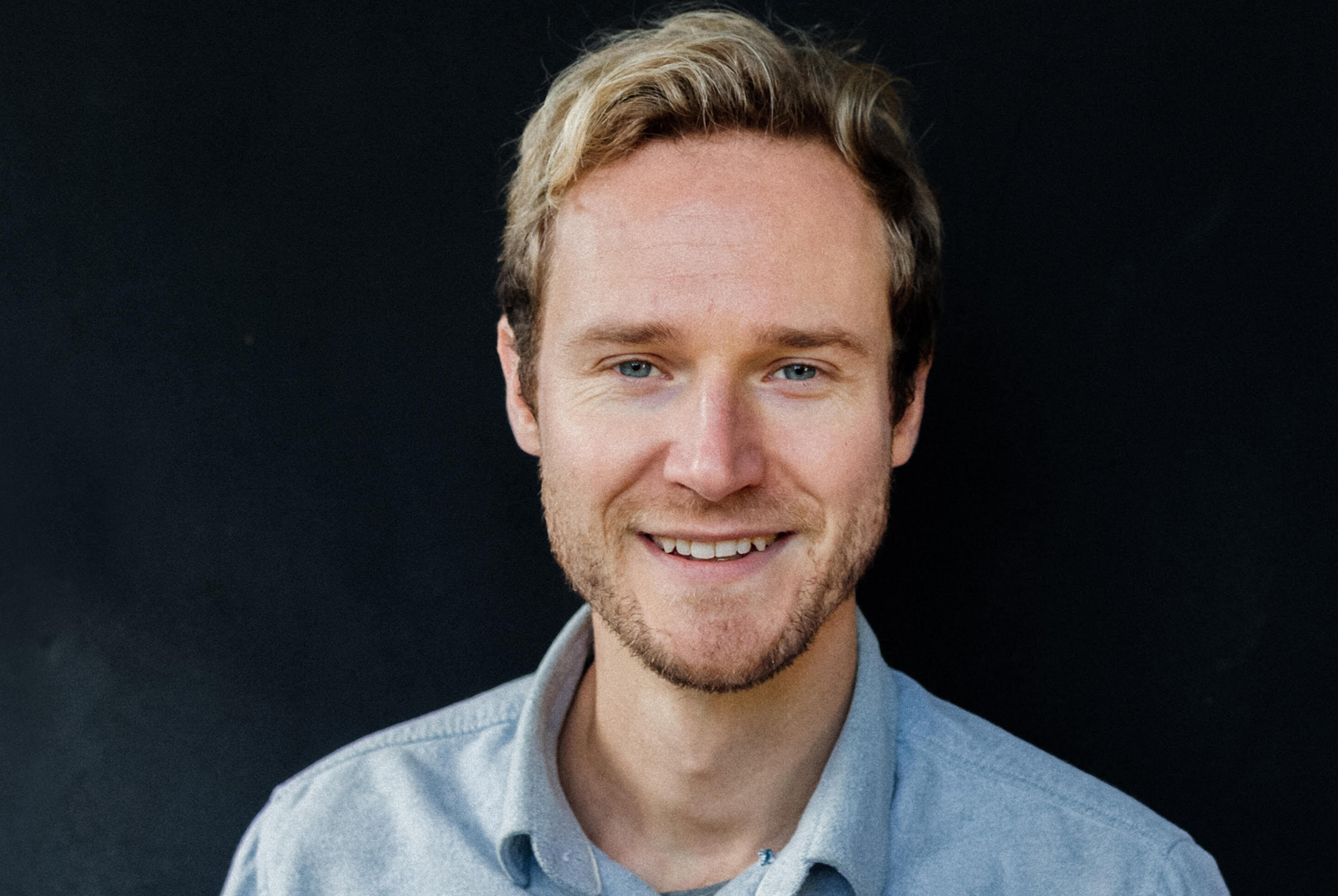 Koos Tervoren glimlachend in blauwe blouse bij zwarte achtergrond