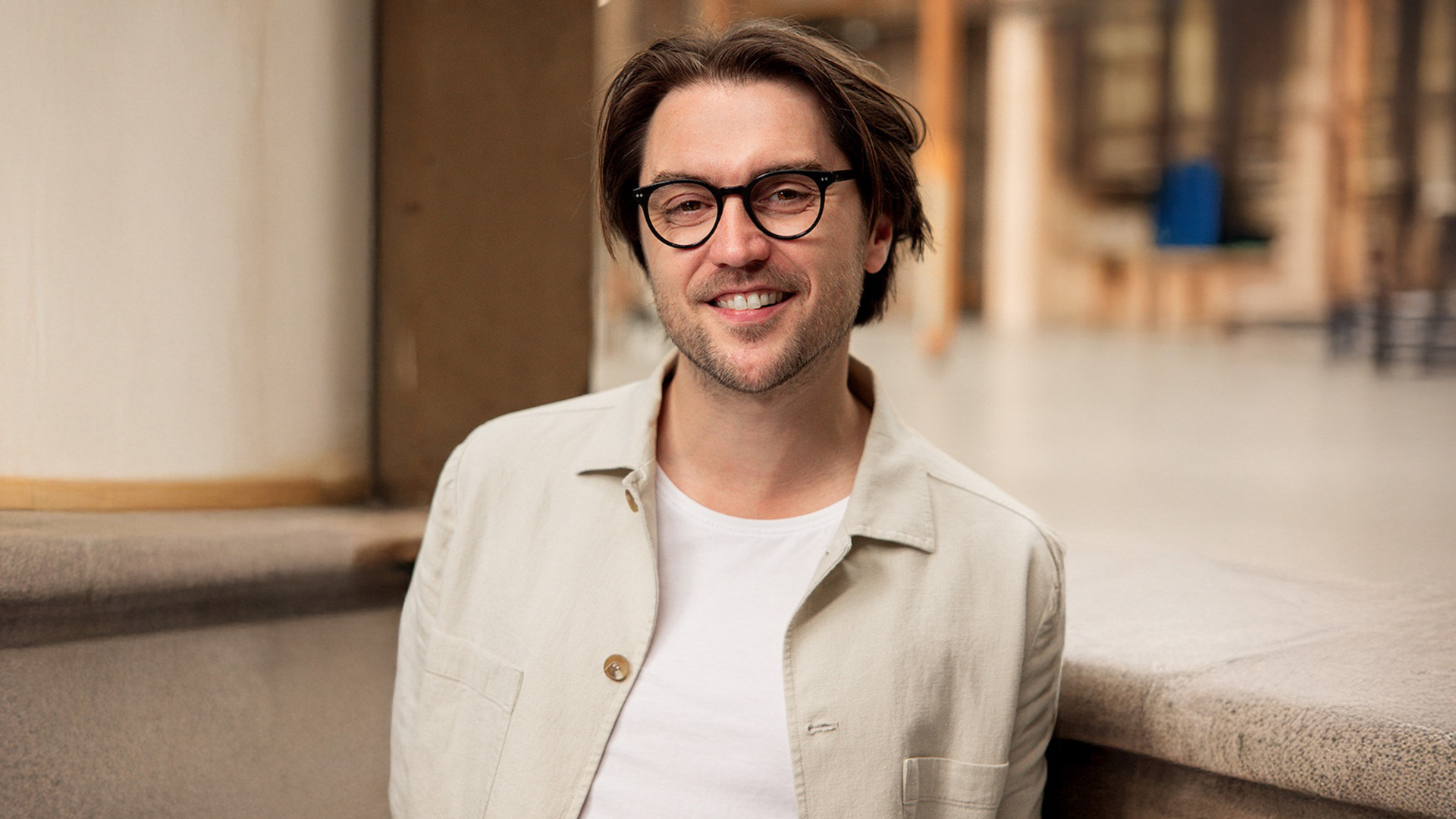 Man met wit shirt aan en een beige jas met een industriele achtergrond