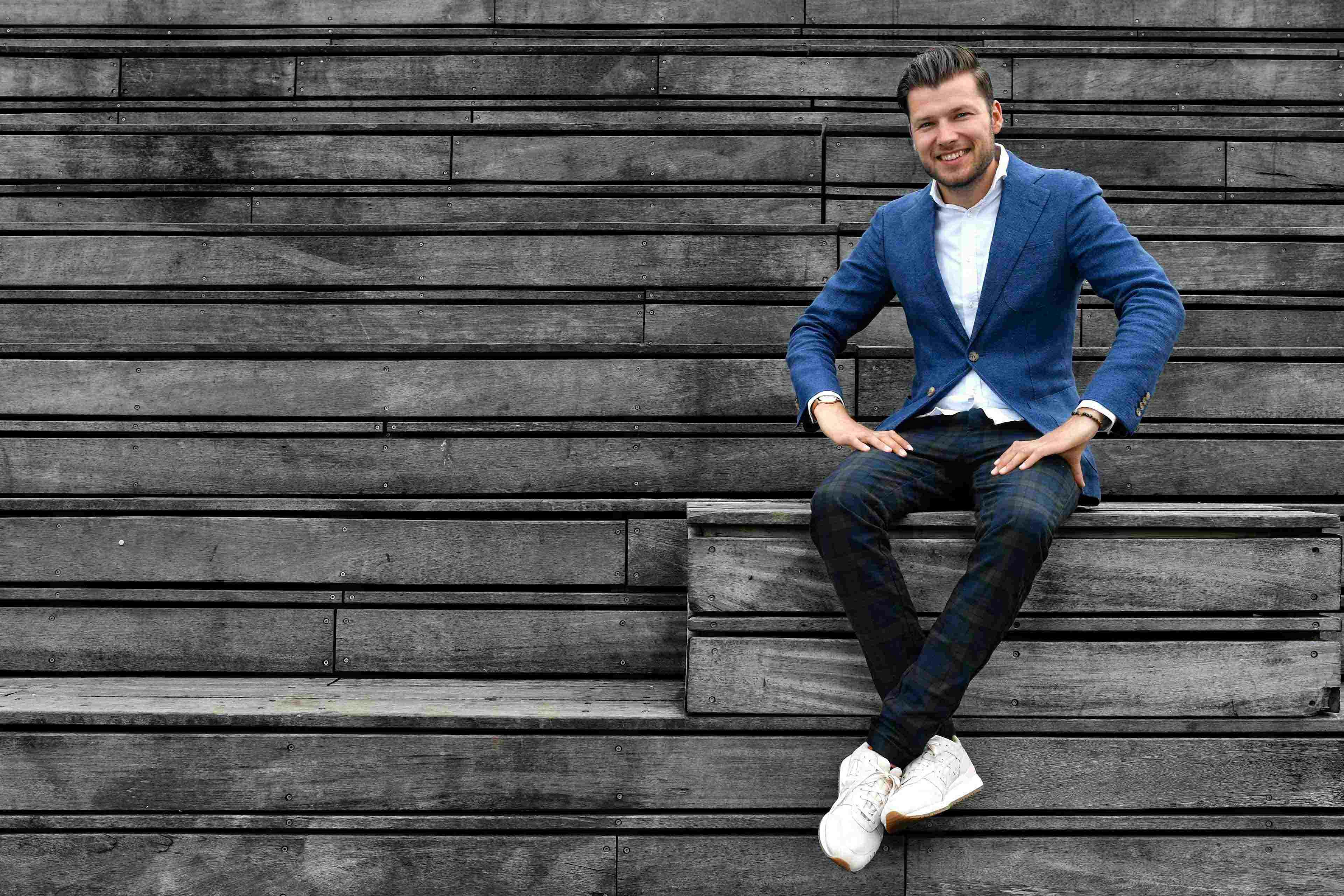 Arjen Banach zittend op houten bankje buiten in blauw pak glimlachend met handen op benen