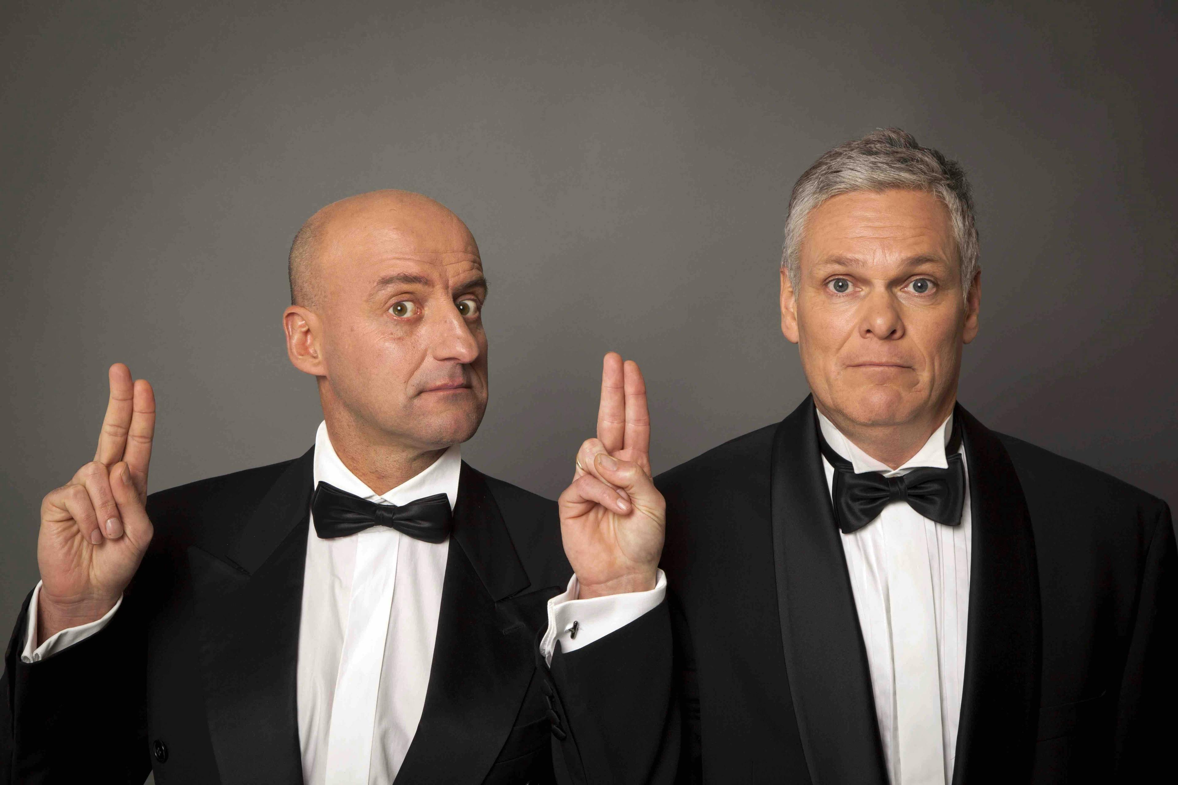 Joep van Deudekom en Rob Urgert in zwart pak die twee vingers opsteken bij grijze achtergrond